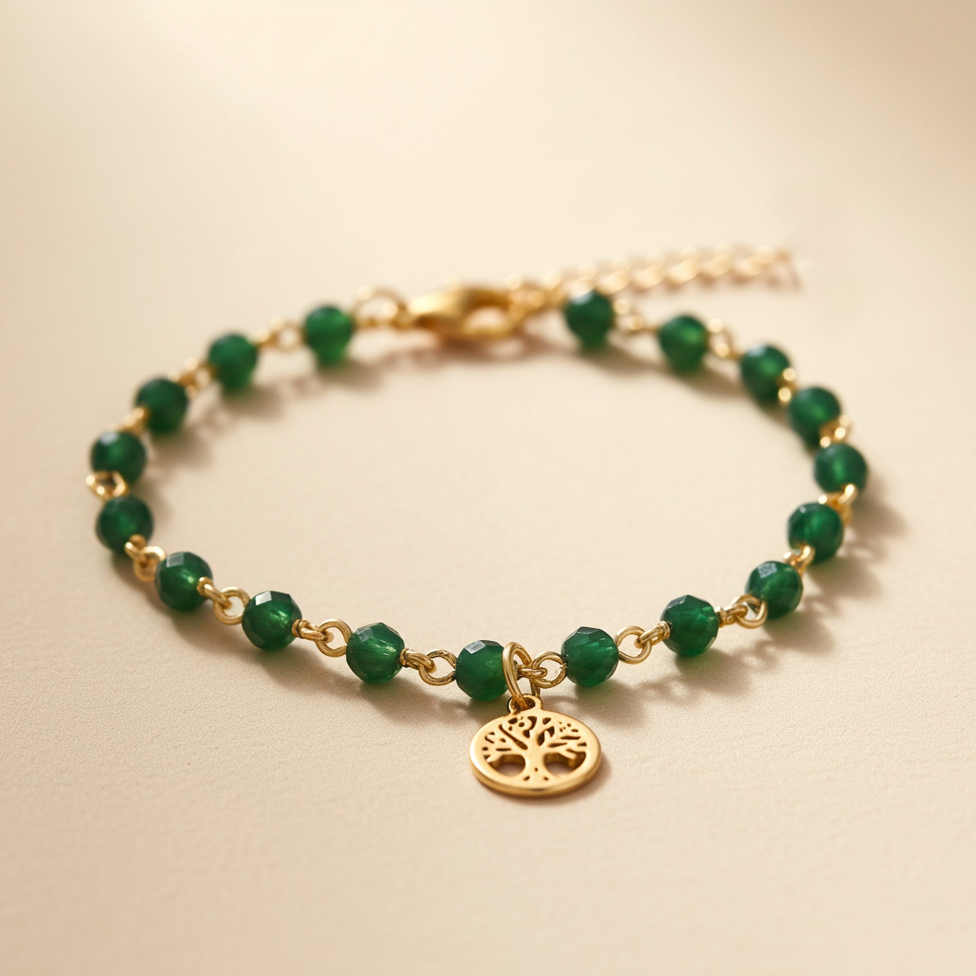 [variant: Vouves Bracelet] Close-up of gold Vouves Olive Tree charm bracelet with green gemstone beads on a warm beige surface
