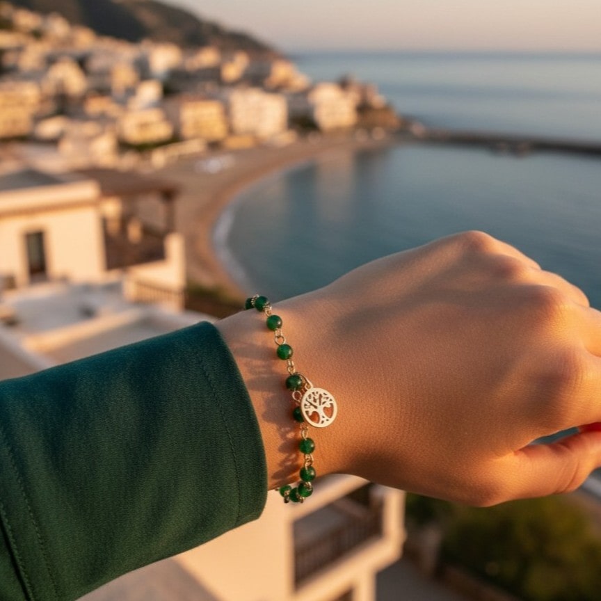 [variant: Vouves Bracelet] Gold Vouves Olive Tree bracelet with green beads worn at sunset overlooking a Mediterranean coastal town