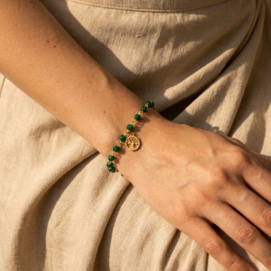 [variant: Vouves Bracelet] Gold Vouves Olive Tree charm bracelet with green gemstone beads worn on wrist in soft natural light on linen