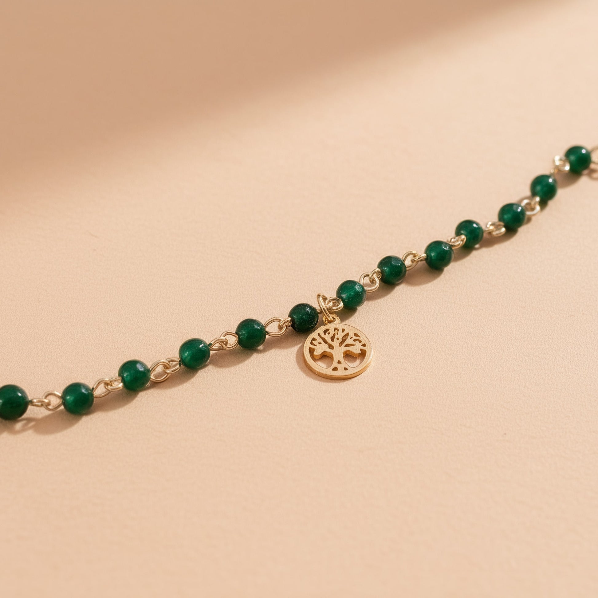 [variant: Vouves Bracelet] Minimal close-up of gold Vouves Olive Tree charm bracelet with green gemstone beads on neutral background