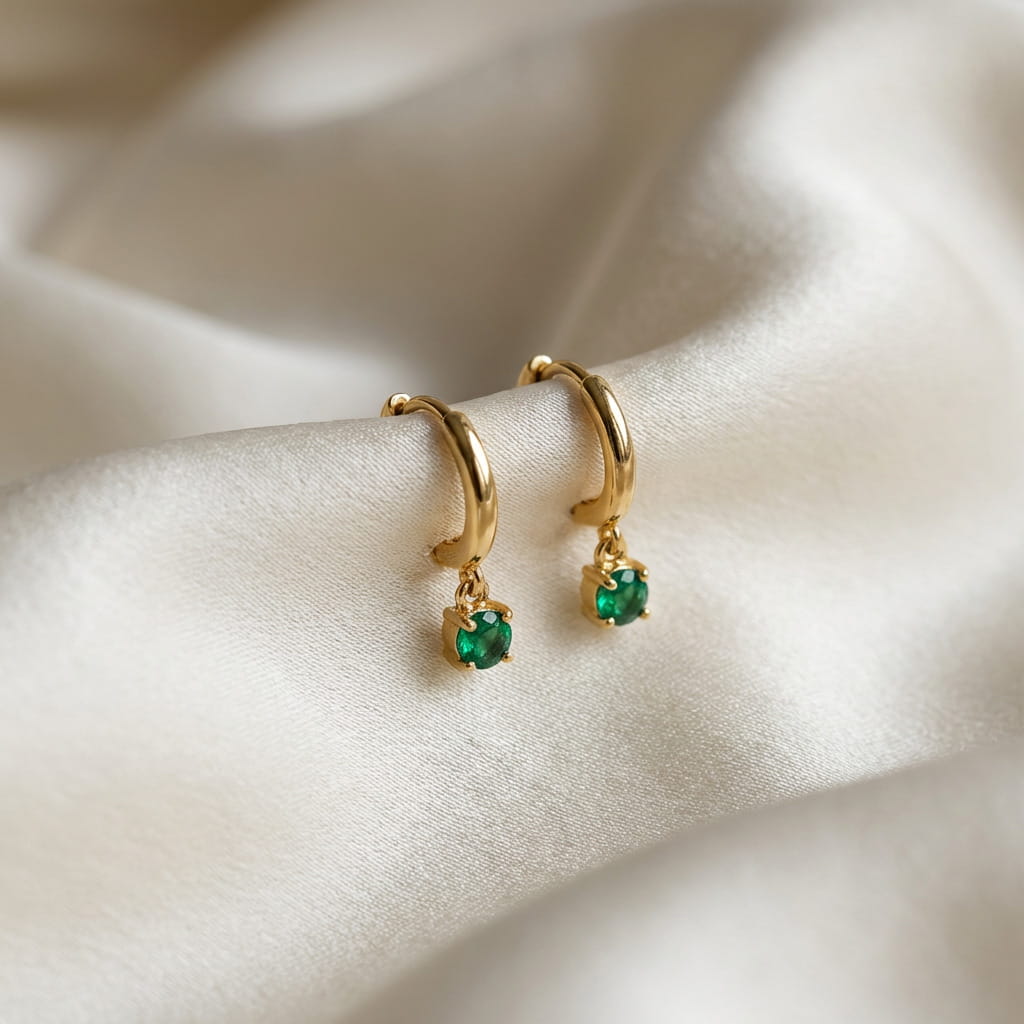 [variant:Vouves Earrings] Gold hoop earrings with green gemstones on a satin surface