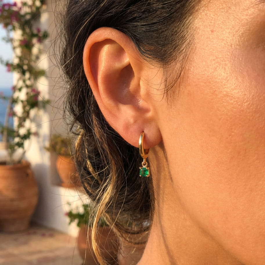 [variant:Vouves Earrings] Close-up of an ear wearing a gold earring with a green gemstone, with a Mediterranean outdoor background.