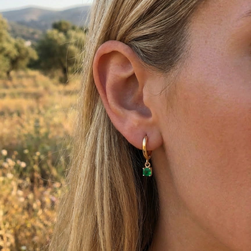 [variant:Full Vouves Collection] Close-up of a person wearing a gold hoop earring with a green gemstone against a blurred natural background.