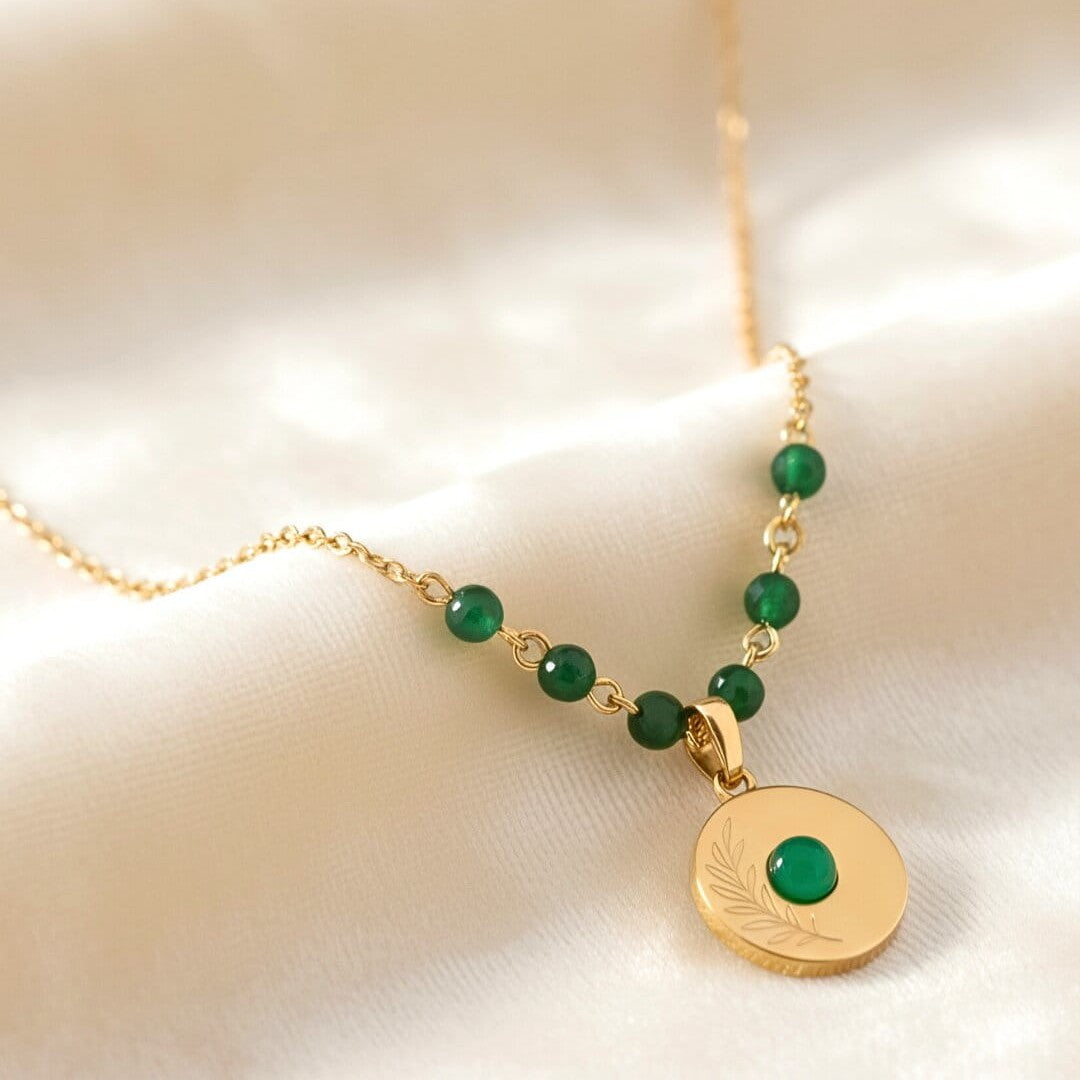 [variant: Vouves Necklace] Gold necklace with green beads and a circular pendant on a beige background