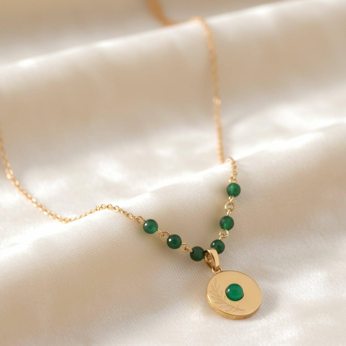 [variant: Vouves Necklace] Gold olive branch pendant necklace with green gemstone beads displayed on soft white fabric