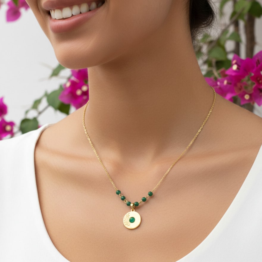 [variant: Vouves Necklace] Gold pendant necklace with green gemstone beads and engraved olive branch worn outdoors near Mediterranean bougainvillea