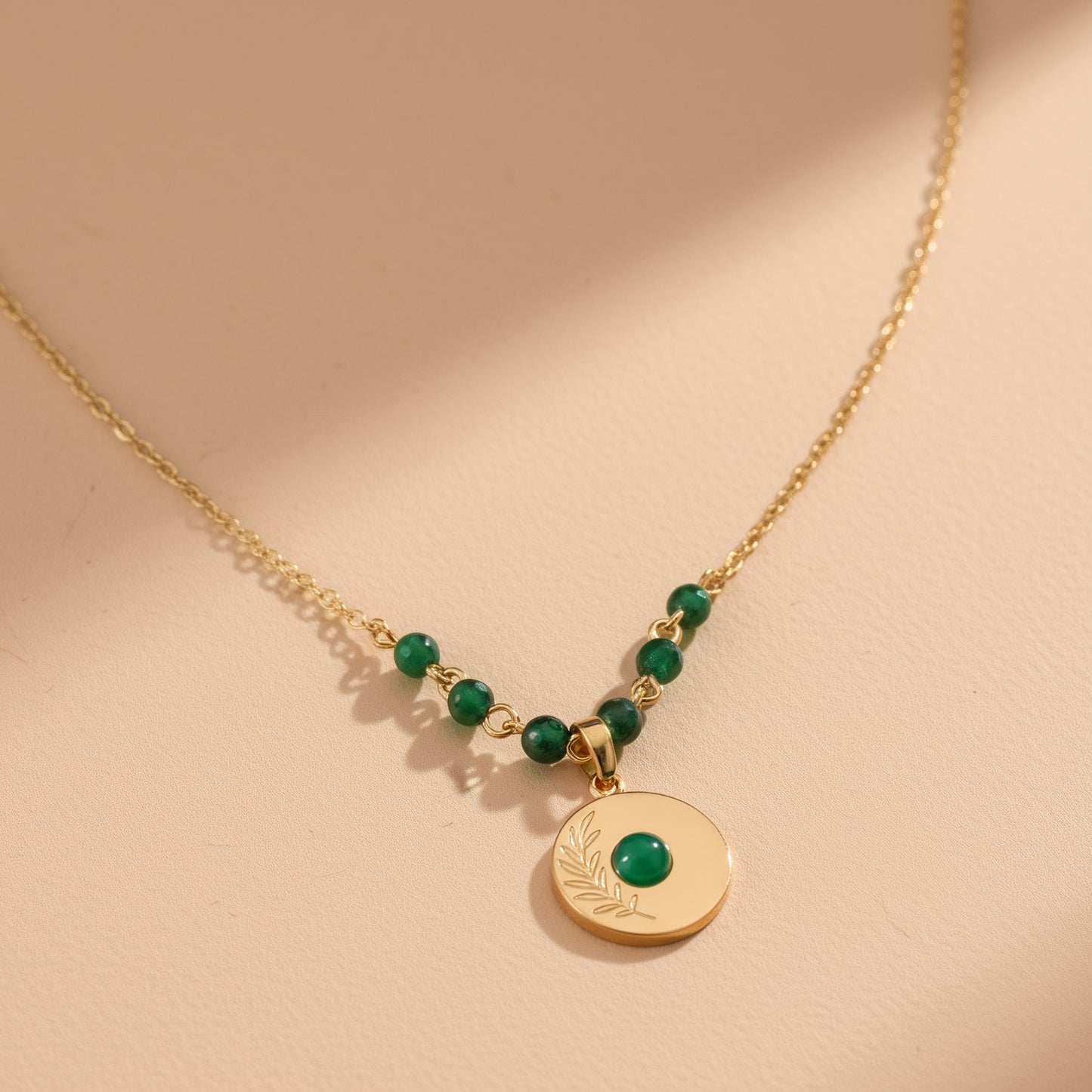 [variant: Vouves Necklace] Gold olive branch engraved pendant necklace with green gemstone beads displayed in warm natural light
