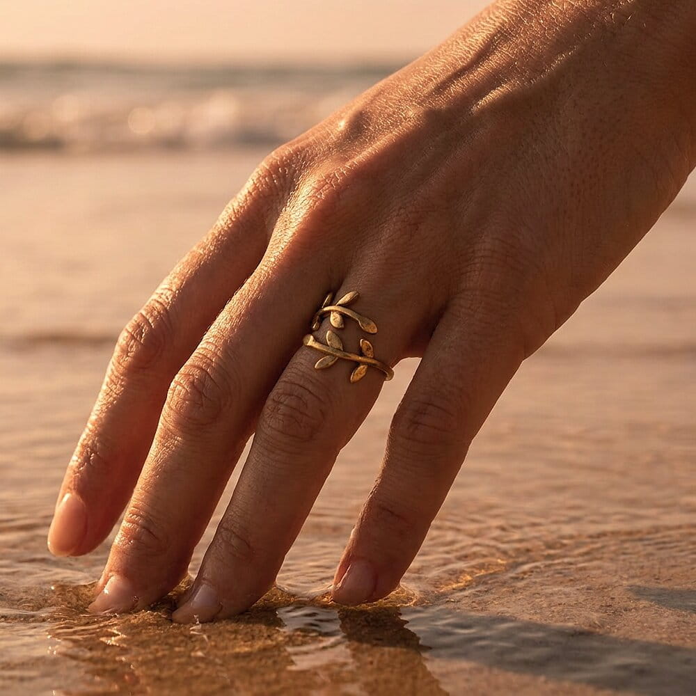 [variant:Vouves Ring] A gold ring with a olive tree branch design worn on a person's hand, which is touching wet sand at a beach during golden hour.