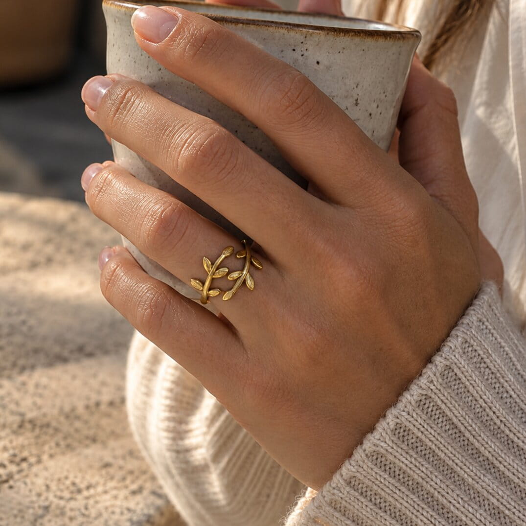 [variant:Vouves Ring] A close-up of a person's hand holding a speckled ceramic mug while wearing a delicate gold leaf branch ring on the ring finger.