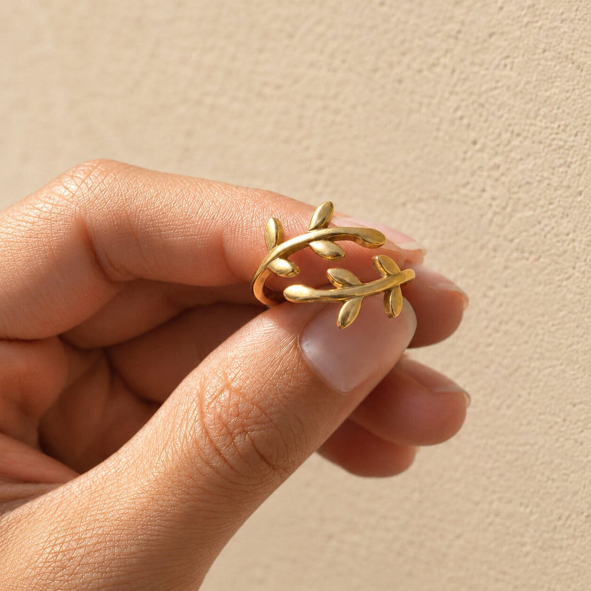 [variant:Vouves Ring] A gold leaf wrap ring being held between fingers against a textured beige background.
