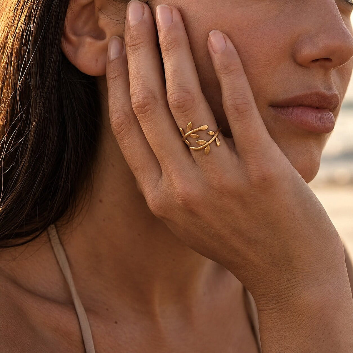[variant:Vouves Ring] A close-up lifestyle shot of a woman wearing a delicate gold ring with a leafy vine design on her ring finger, with her hand resting against her cheek.